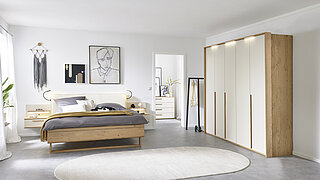 Vorschlag 701, Balkeneiche furniert, Absetzung Lack Sand, bestehend aus Drehtürenschrank 6-türig, 3 Griffstangen in Eiche massiv, ca. B 299, H 229, T 60 cm, Doppelbett in Luxushöhe, ca. B 189, H 105, T 222 cm, Liegefläche ca. 180 x 200 cm, BSH 48,6 cm, Polsterkopfteil "Luna" in Stoff Maranta Sand, 100 % Polyester, Fußteil mit Lisene, unten abgerundet und Kufe in Eiche, 2 Hängekonsolen mit je 1 Schubkasten, jeweils ca. B 60, H 23, T 46 cm. Zubehör: Passepartout ohne Beleuchtung (903004); 3er-Set LED-Stabbeleuchtung (90353); Paneelaufsatz links und rechts mit Holzboden (20216, 20226); indirekte LED-Holzbodenbeleuchtung (80027); 2er-Set LED-Flex-Leuchte Anthrazit, USB-Anschluss (80033).