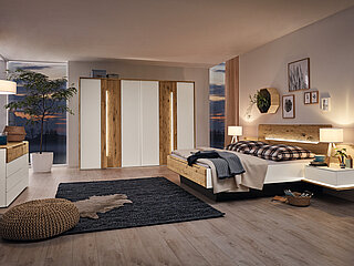 Schlafzimmermöbel Jovanna, Vorschlag 2: Drehtürenschrank 6-trg., 2 Designtüren in Absetzung, Bett mit Holzkopfteil und Fußteilblende, Ansatzregal links und rechts mit Ablageboden (Zubehör: Aufsatzschubkasten links und rechts)B 298,6, H 222,3, T 64 cm (Schrank), 180 x 200 cm (Bett), je B 72,3, H 59, T 67,5 cm (Ansatzpaneel), B 58,5, H 22, T 45,1 cm (Aufsatzschubkasten)Lack weiß/Absetzungen Wildeiche massiv